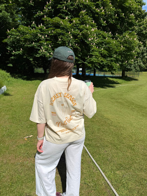 A person standing outdoors on a grassy lawn wearing a cream-colored t-shirt with orange graphic text that reads 'WICKET SEASON' and light gray sweatpants, viewed from behind.