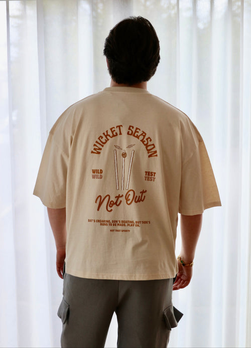 A person viewed from behind wearing a beige oversized cricket inspired T-shirt with a large brown graphic print on the back reading 'WICKET SEASON Not Out' and 'WILD TEST TEST', paired with gray cargo pants in front of white sheer curtains.

